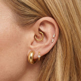 Close-up of a woman’s ear styled with multiple gold earrings. The ear features a thick, smooth gold huggie in the lobe, a smaller gold hoop with a tiny star-shaped diamond charm beside it, a textured gold hoop in the upper cartilage, and two small gold studs with delicate diamonds placed vertically along the helix. The model’s hair is tucked behind the ear, highlighting the curated gold stack.