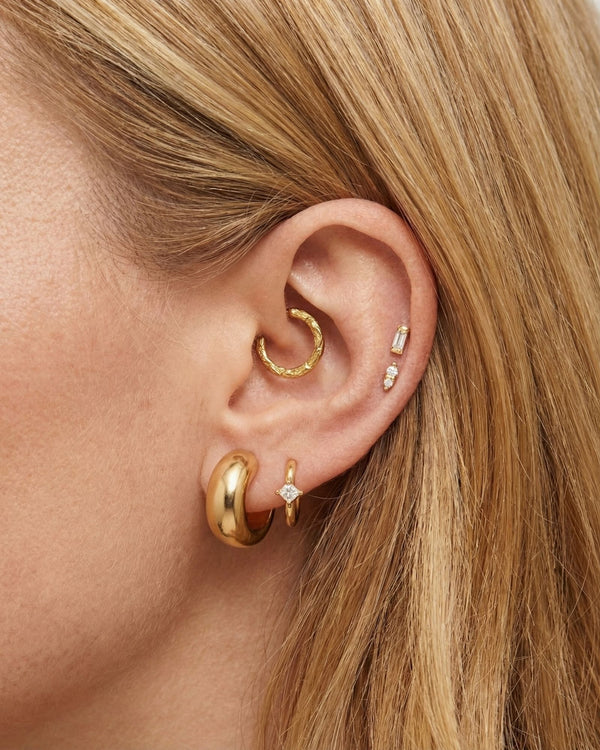Close-up of a woman’s ear styled with multiple gold earrings. The ear features a thick, smooth gold huggie in the lobe, a smaller gold hoop with a tiny star-shaped diamond charm beside it, a textured gold hoop in the upper cartilage, and two small gold studs with delicate diamonds placed vertically along the helix. The model’s hair is tucked behind the ear, highlighting the curated gold stack.