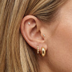 Close-up side profile of a woman’s ear wearing a layered gold earring stack, including a polished gold hoop in the lobe, a slim gold hoop with small pearl accents, and delicate gold studs in the cartilage and inner ear. The image clearly shows the warm gold tones, varied textures, and placement across multiple piercings. Designed for women who enjoy curated ear styling, this image helps customers visualise how to mix gold hoops and stud earrings to create a balanced, refined everyday ear stack.