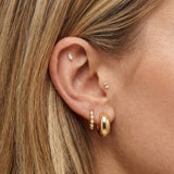 Close-up side profile of a woman’s ear wearing a layered gold earring stack, including a polished gold hoop in the lobe, a slim gold hoop with small pearl accents, and delicate gold studs in the cartilage and inner ear. The image clearly shows the warm gold tones, varied textures, and placement across multiple piercings. Designed for women who enjoy curated ear styling, this image helps customers visualise how to mix gold hoops and stud earrings to create a balanced, refined everyday ear stack.