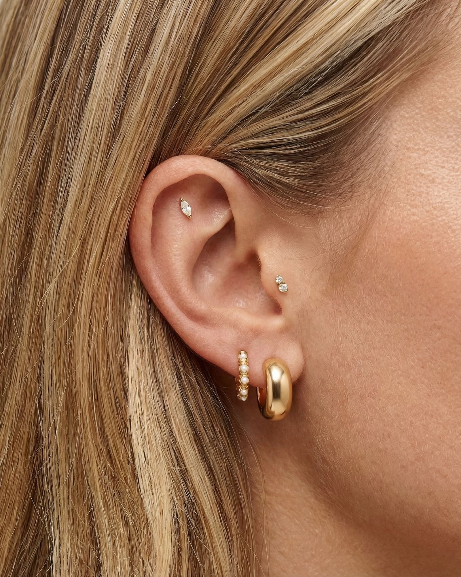 Close-up side profile of a woman’s ear wearing a layered gold earring stack, including a polished gold hoop in the lobe, a slim gold hoop with small pearl accents, and delicate gold studs in the cartilage and inner ear. The image clearly shows the warm gold tones, varied textures, and placement across multiple piercings. Designed for women who enjoy curated ear styling, this image helps customers visualise how to mix gold hoops and stud earrings to create a balanced, refined everyday ear stack.