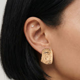 Close-up side profile of a woman’s ear wearing a gold statement stud earring with an abstract, sculptural rectangular form, paired with a small marquise-style cartilage stud. Created for women who prefer modern gold jewellery with a strong design presence, this image shows how the gold stud sits on the ear and how it can be styled with additional piercings for a polished, contemporary look.