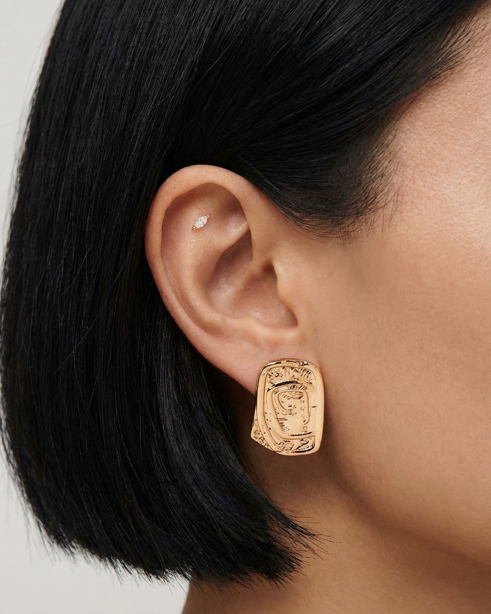 Close-up side profile of a woman’s ear wearing a gold statement stud earring with an abstract, sculptural rectangular form, paired with a small marquise-style cartilage stud. Created for women who prefer modern gold jewellery with a strong design presence, this image shows how the gold stud sits on the ear and how it can be styled with additional piercings for a polished, contemporary look.