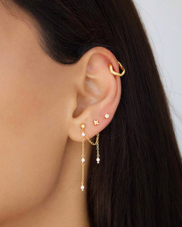 Close-up of a woman wearing gold chain drop earrings with freshwater pearls and a cubic zirconia stud, styled in a layered gold ear stack.