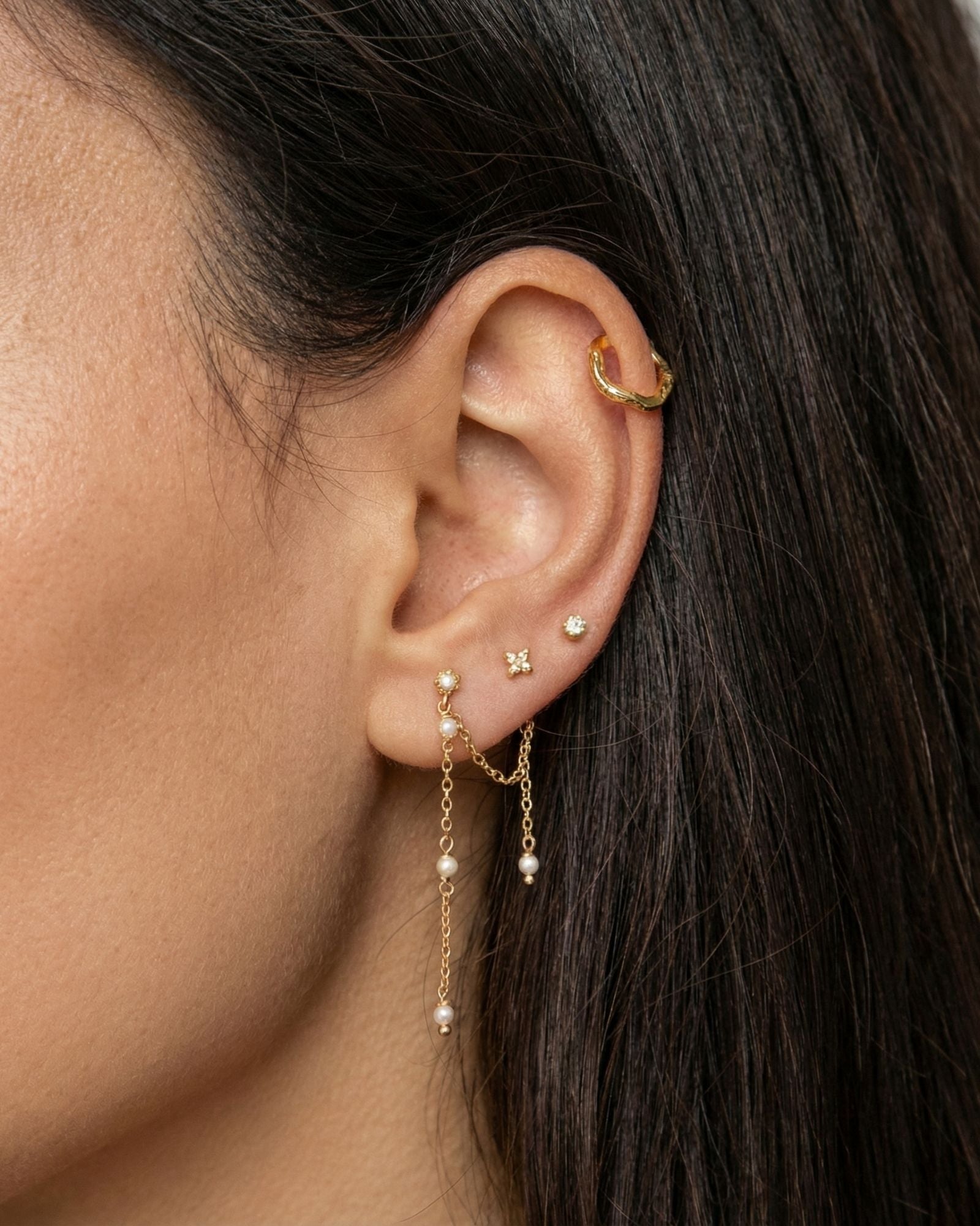 Close-up side profile of a woman’s ear wearing a layered gold ear stack, featuring a textured gold ear cuff on the upper cartilage, delicate gold stud earrings, and a fine gold chain drop earring accented with small pearls. The image clearly shows the soft movement of the chain, the contrast between polished gold and pearl details, and the placement across multiple piercings. Designed for women who enjoy elegant, feminine ear styling, this image helps customers visualise how to combine gold studs, cuffs, an