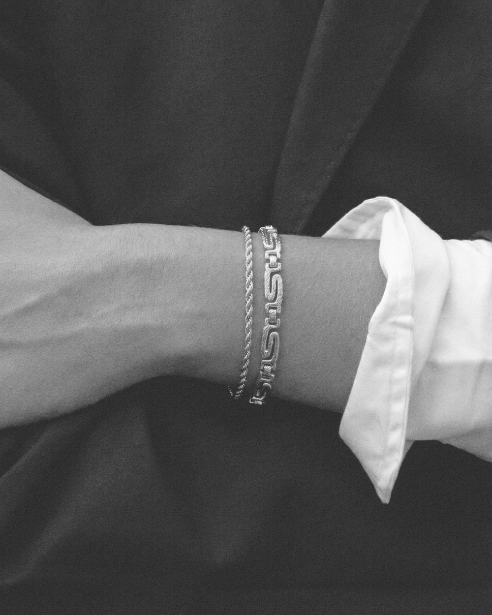 Black and white close-up of a woman’s wrist wearing two silver bracelets: a fine rope chain bracelet and a bold geometric link bracelet, styled together against a dark fabric background with a white shirt cuff visible. The image clearly shows the contrast between the delicate chain and the structured link design, as well as how the bracelets sit when layered on the wrist. Designed for women who enjoy stacking silver bracelets, this image helps customers visualise layering options, scale, and how mixing mini
