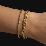 Close-up of a woman’s wrist wearing two gold bracelets layered together: a slim gold rope chain bracelet and a bold textured geometric link bracelet, photographed against a black background. The image clearly shows the contrast between the delicate rope design and the structured statement links, as well as the warm gold tone and polished finish. Designed for women who enjoy layering gold bracelets, this image helps customers visualise stacking options, scale, and how mixing fine and bold gold bracelets 