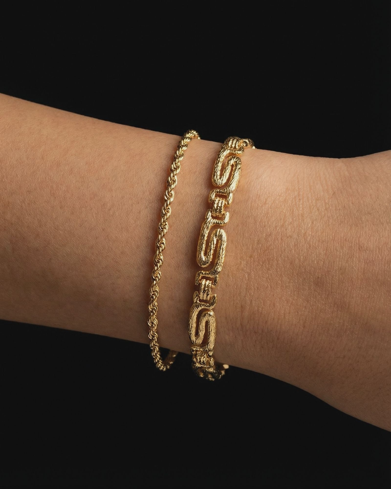 Close-up of a woman’s wrist wearing two gold bracelets layered together: a slim gold rope chain bracelet and a bold textured geometric link bracelet, photographed against a black background. The image clearly shows the contrast between the delicate rope design and the structured statement links, as well as the warm gold tone and polished finish. Designed for women who enjoy layering gold bracelets, this image helps customers visualise stacking options, scale, and how mixing fine and bold gold bracelets 