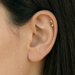 Close-up of a woman’s ear wearing a delicate gold flat back cartilage stud set with a small green gemstone and two clear accent stones, positioned in the upper helix. Designed for women seeking minimalist gold cartilage earrings, this image highlights a secure flat back ear piercing that offers comfort for sensitive ears, new or healed piercings, and everyday wear while adding a subtle pop of colour to a curated ear stack.