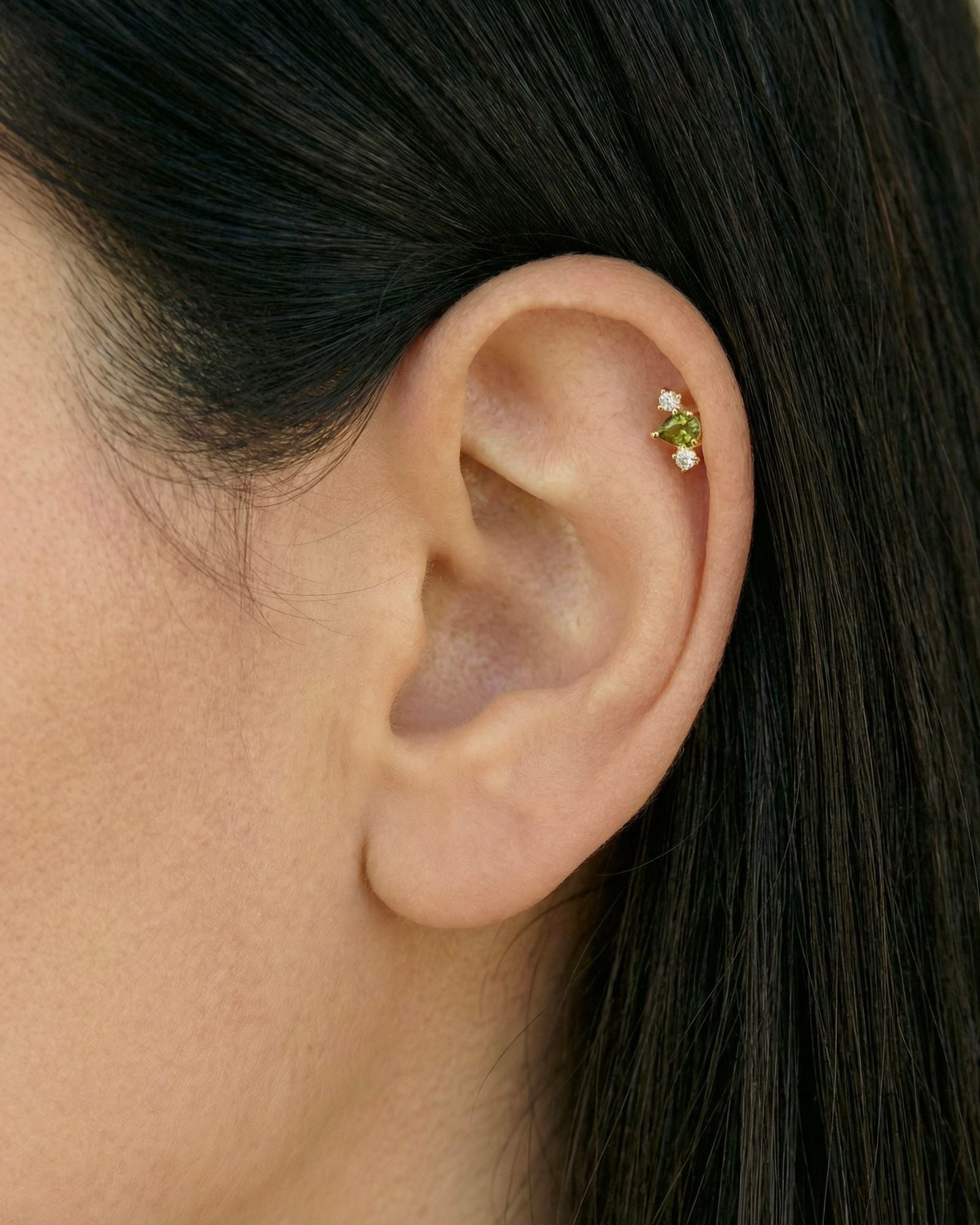 Close-up of a woman’s ear wearing a delicate gold flat back cartilage stud set with a small green gemstone and two clear accent stones, positioned in the upper helix. Designed for women seeking minimalist gold cartilage earrings, this image highlights a secure flat back ear piercing that offers comfort for sensitive ears, new or healed piercings, and everyday wear while adding a subtle pop of colour to a curated ear stack.