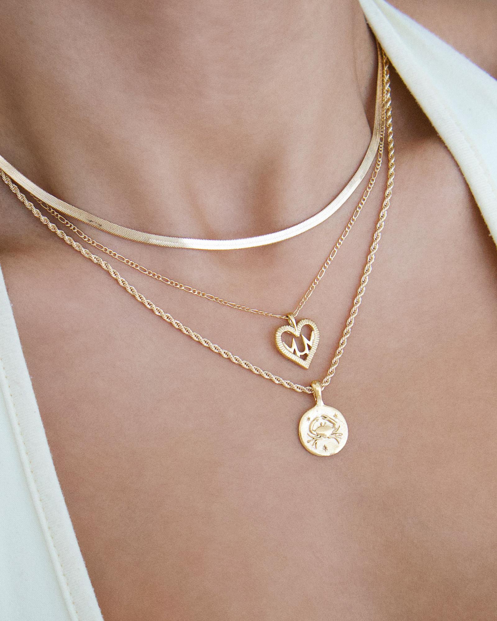 Close-up lifestyle image of layered gold necklaces featuring a flat gold snake chain worn closest to the neck, paired with two fine gold chains with heart and round coin pendants. The jewellery is styled on bare skin beneath a light top, showing how a snake chain anchors a layered necklace look. Designed for women seeking versatile gold jewellery and inspiration for elegant layering, helping solve the challenge of building a balanced, everyday necklace stack with statement and minimal pieces.