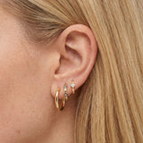 Close-up of a woman’s ear wearing a curated stack of small gold hoop and huggie earrings, including a smooth gold hoop, a slim huggie with blue and clear cubic zirconia accents, and a minimalist gold huggie with a square-cut clear crystal. The image shows layered gold huggie earrings styled on multiple lobe piercings, designed for women who want a refined, modern ear stack with subtle colour and sparkle. Ideal for everyday wear, work, or elevated casual styling, this ear stack helps solve the need for versa