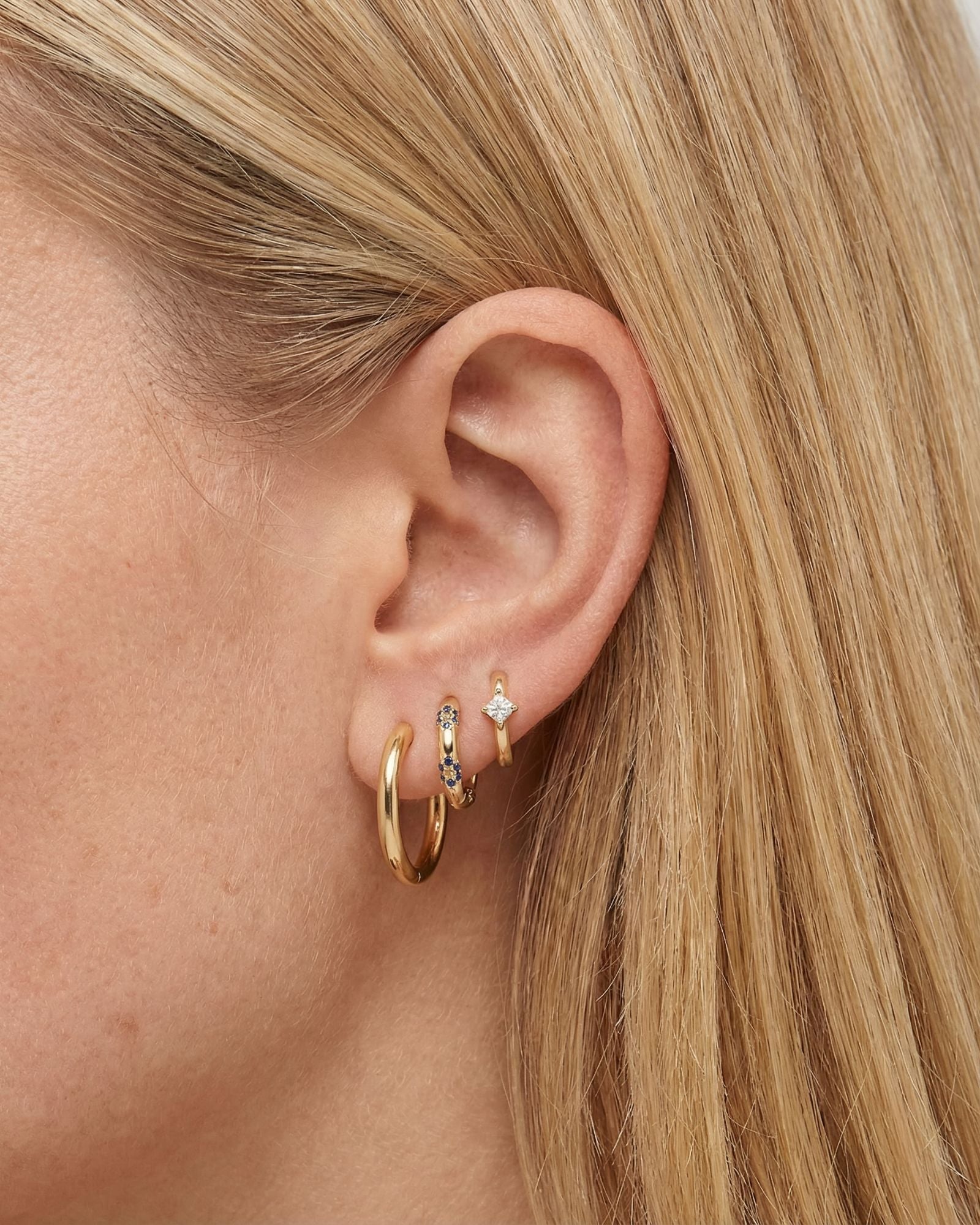 Close-up product photo of a woman’s ear with fair skin and straight blonde hair, styled with layered gold earrings across multiple lobe piercings. The ear features a smooth medium-sized gold hoop in the first piercing, a slim gold huggie set with small blue stones in the second piercing, and a delicate gold huggie with a clear stone set in a star-shaped design in the third piercing. This image is for customers looking for elegant gold earrings that add a subtle pop of colour while remaining suitable for eve