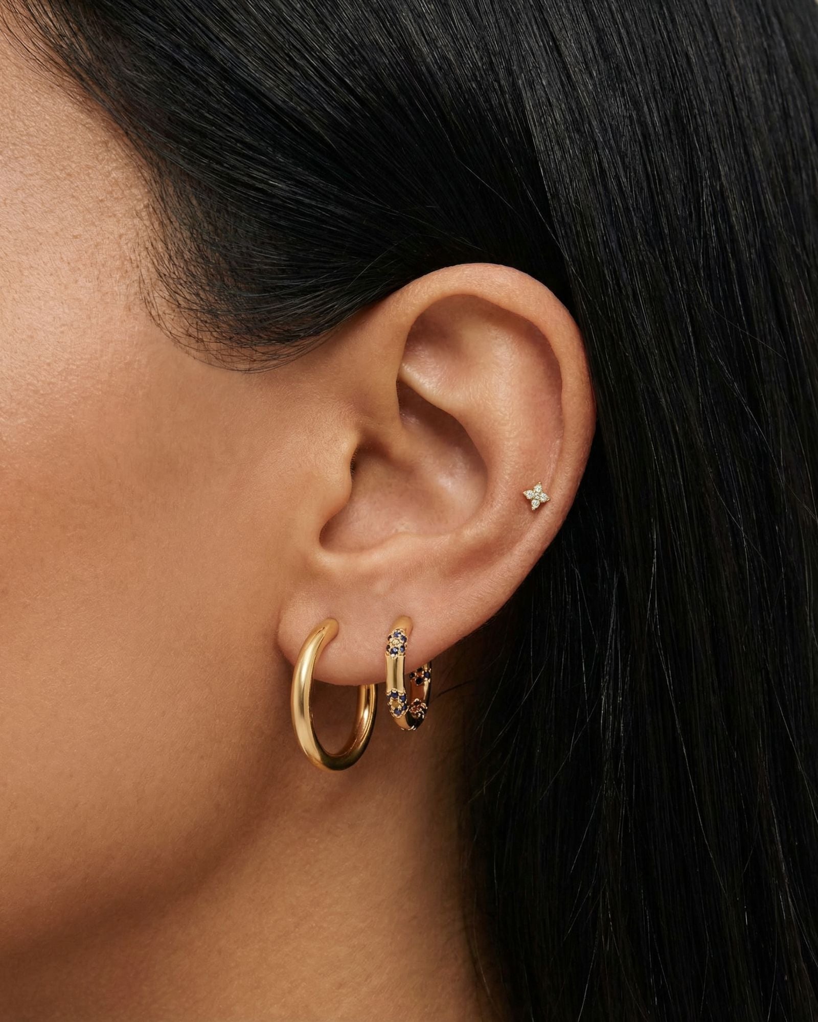 Close-up of a woman’s ear styled with a gold ear stack featuring a smooth gold hoop, a blue crystal huggie hoop, and a tiny star-shaped flat back stud in the upper cartilage. This image is for women looking for flat back earrings, cartilage piercings, and minimalist gold jewelry, showing how small, low-profile studs and hoops can be layered comfortably for everyday wear, sensitive ears, and a clean, modern ear stack.