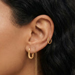 Close-up product photo of a woman’s ear with warm medium-brown skin and dark natural hair, styled with coordinated gold earrings. The ear features a small textured gold hoop in the upper cartilage, a smooth gold huggie hoop in the lobe, and a delicate gold chain drop earring with double slim bar pendants. This image is for customers seeking versatile gold earrings that work across multiple piercings and suit daily wear. It shows how minimalist gold jewellery can create a cohesive, elevated ear stack while s