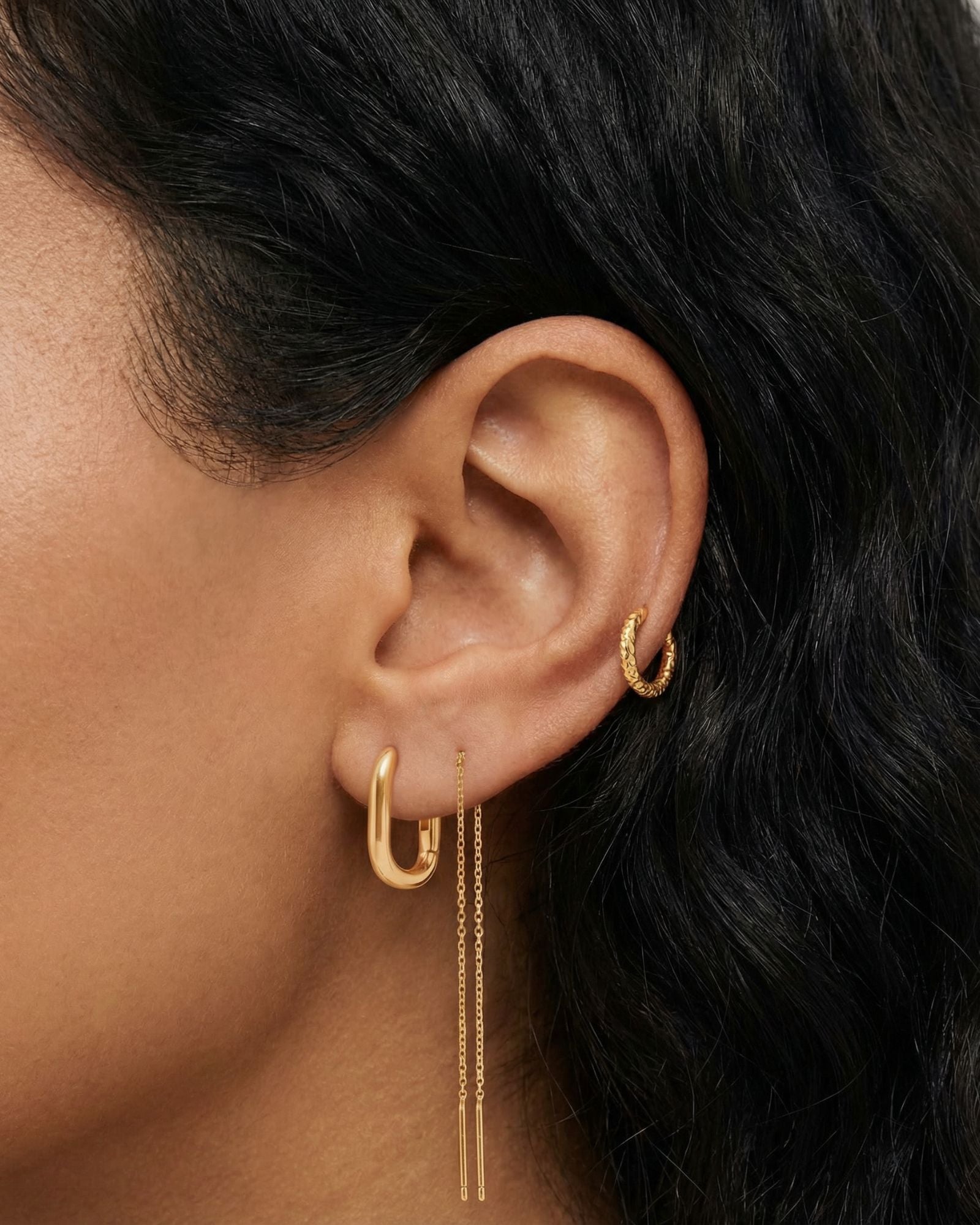 Close-up product photo of a woman’s ear with warm medium-brown skin and dark natural hair, styled with coordinated gold earrings. The ear features a small textured gold hoop in the upper cartilage, a smooth gold huggie hoop in the lobe, and a delicate gold chain drop earring with double slim bar pendants. This image is for customers seeking versatile gold earrings that work across multiple piercings and suit daily wear. It shows how minimalist gold jewellery can create a cohesive, elevated ear stack while s