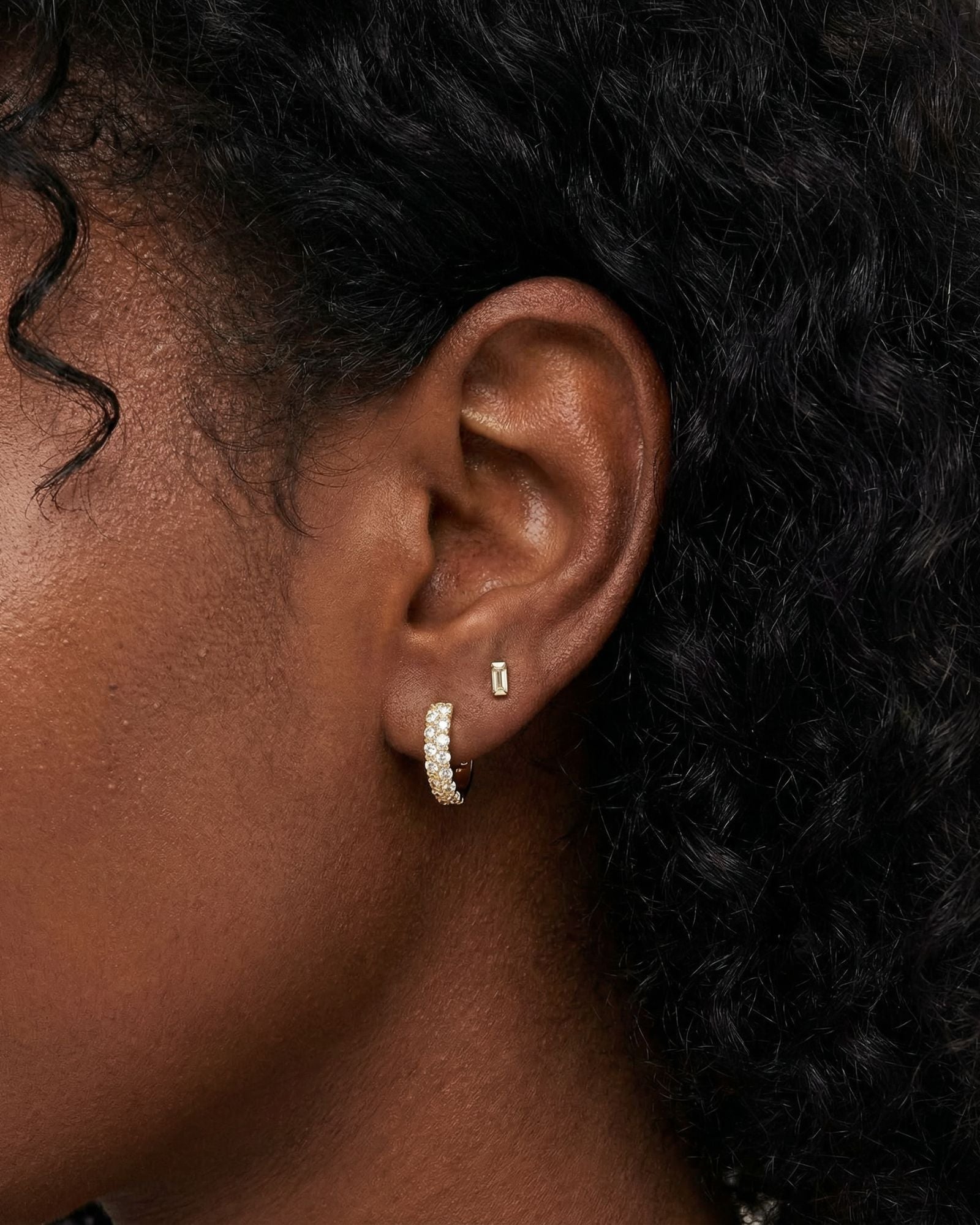 Close-up lifestyle image of a woman wearing Tia crystal gold hoop earrings on the lobe, styled with small gold stud earrings  and minimalist bar stud. Ideal for women looking for small gold crystal hoops that add refined sparkle, are lightweight for sensitive ears, and create an effortless, everyday ear stack with subtle shine.
