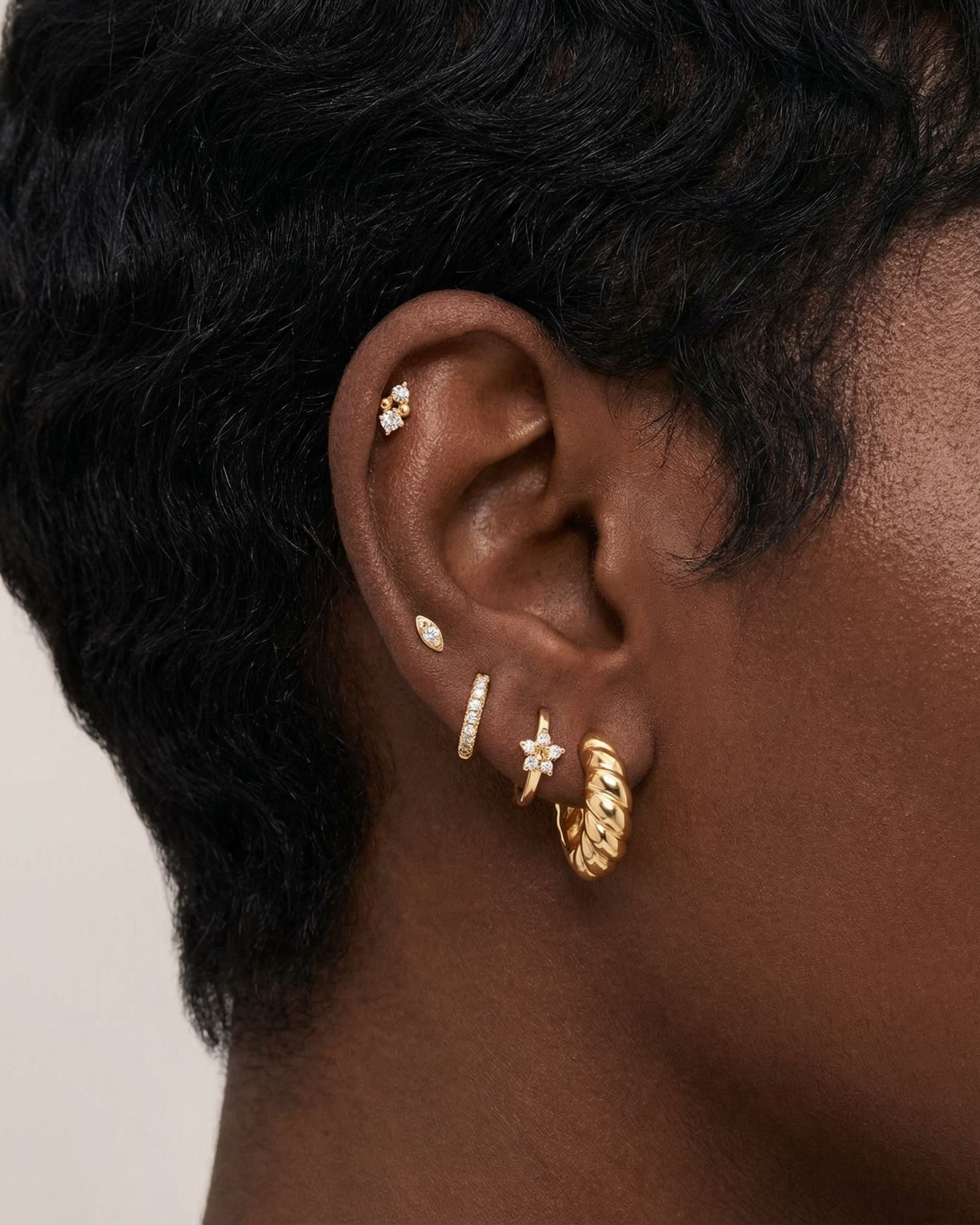 Close-up side profile of a blonde woman’s ear wearing a curated gold ear stack, featuring a twisted gold hoop earring, a slim pavé huggie, small marquise and round crystal studs, and a delicate cartilage stud. Designed for women with multiple piercings, this image helps customers visualise scale, placement, and layering of gold hoops and stud earrings to create a refined, cohesive everyday ear stack.