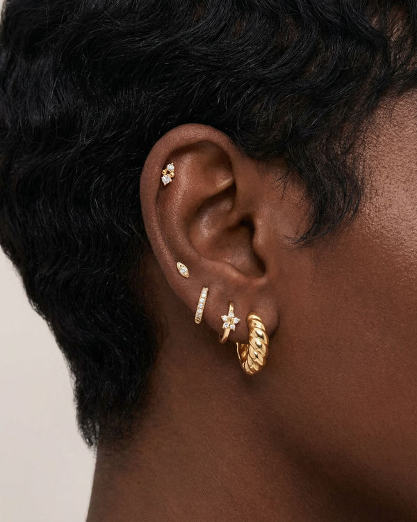 Close-up side profile of a blonde woman’s ear wearing a curated gold ear stack, featuring a twisted gold hoop earring, a slim pavé huggie, small marquise and round crystal studs, and a delicate cartilage stud. Designed for women with multiple piercings, this image helps customers visualise scale, placement, and layering of gold hoops and stud earrings to create a refined, cohesive everyday ear stack.