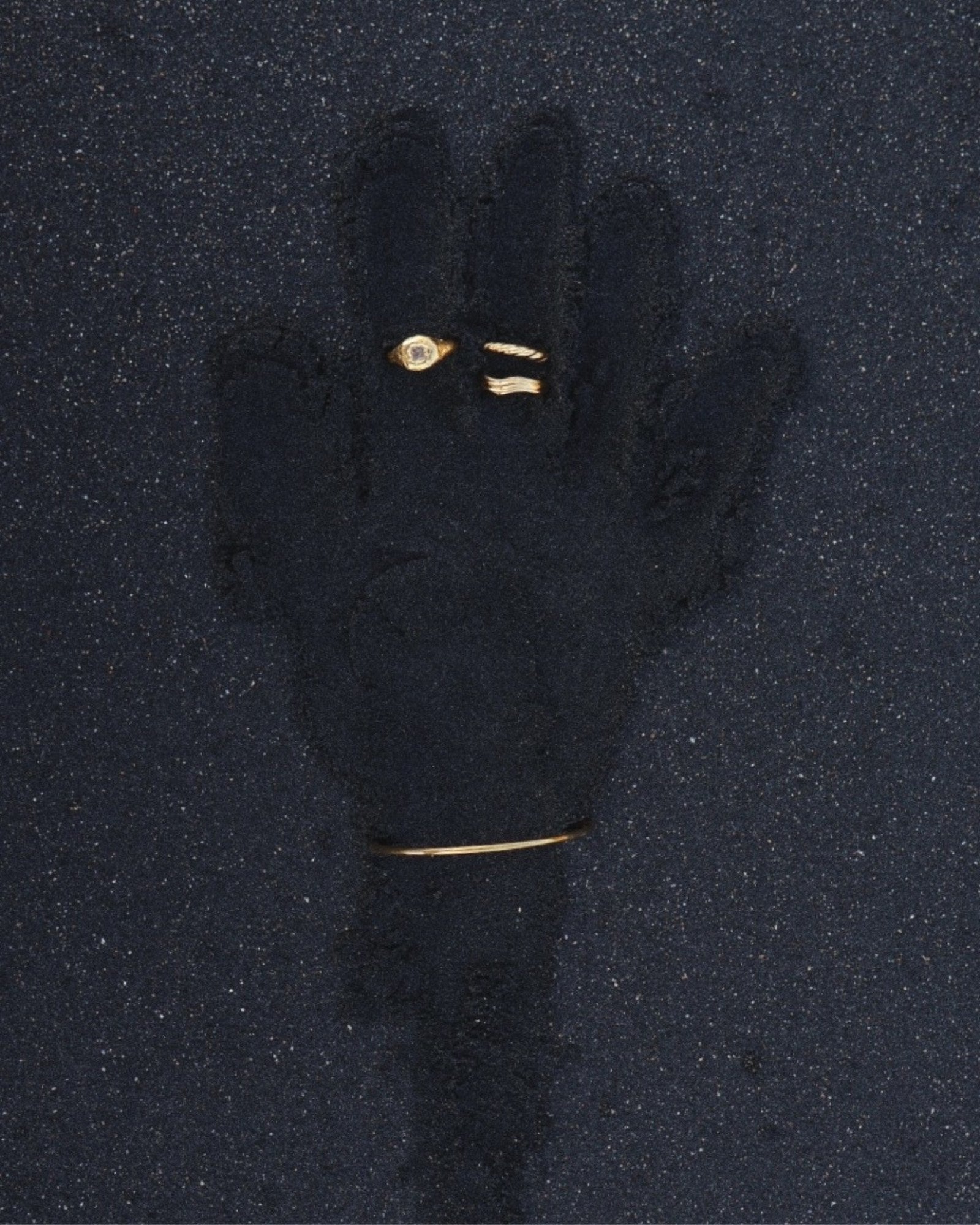 Flat lay of minimalist gold jewellery arranged on dark textured sand, featuring a slim gold bangle bracelet, a delicate gold ring with a clear stone, and two small gold bands. Designed for women who prefer understated, everyday gold jewellery, this image highlights a cohesive, minimalist jewellery set ideal for layering and stacking. The photo helps customers visualise how simple gold bangles and rings can be styled together for a refined, modern look with low-maintenance elegance.