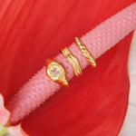 Product image showing three gold rings styled together on a textured pink stem against a red floral background: a small organic gold signet ring with a clear stone, a minimalist gold wave ring, and a slim gold band. Designed for those searching for gold stacking rings, minimalist gold jewellery, and everyday statement rings, this image highlights versatile gold rings that solve the need for easy layering, cohesive styling, and refined everyday wear with a modern, organic aesthetic.