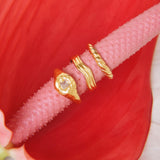 Product image showing three gold rings styled together on a textured pink stem against a red floral background: a small organic gold signet ring with a clear stone, a minimalist gold wave ring, and a slim gold band. Designed for those searching for gold stacking rings, minimalist gold jewellery, and everyday statement rings, this image highlights versatile gold rings that solve the need for easy layering, cohesive styling, and refined everyday wear with a modern, organic aesthetic.