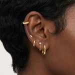Close-up product photo of a woman’s ear with deep brown skin and natural textured hair, styled with layered gold earrings across multiple piercings. The ear features a textured gold ear cuff along the upper cartilage, a small marquise-shaped stone stud, a slim textured gold huggie, a delicate gold chain drop earring with a small round stone, and a smooth gold huggie hoop in the lobe. This image is for customers looking for versatile gold earrings that work across multiple piercings and ear placements. It he