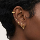 Close-up product photo of a woman’s ear with deep brown skin and natural textured hair, styled with layered gold earrings across multiple piercings. The ear features a textured gold ear cuff along the upper cartilage, a small marquise-shaped stone stud, a slim textured gold huggie, a delicate gold chain drop earring with a small round stone, and a smooth gold huggie hoop in the lobe. This image is for customers looking for versatile gold earrings that work across multiple piercings and ear placements. It he