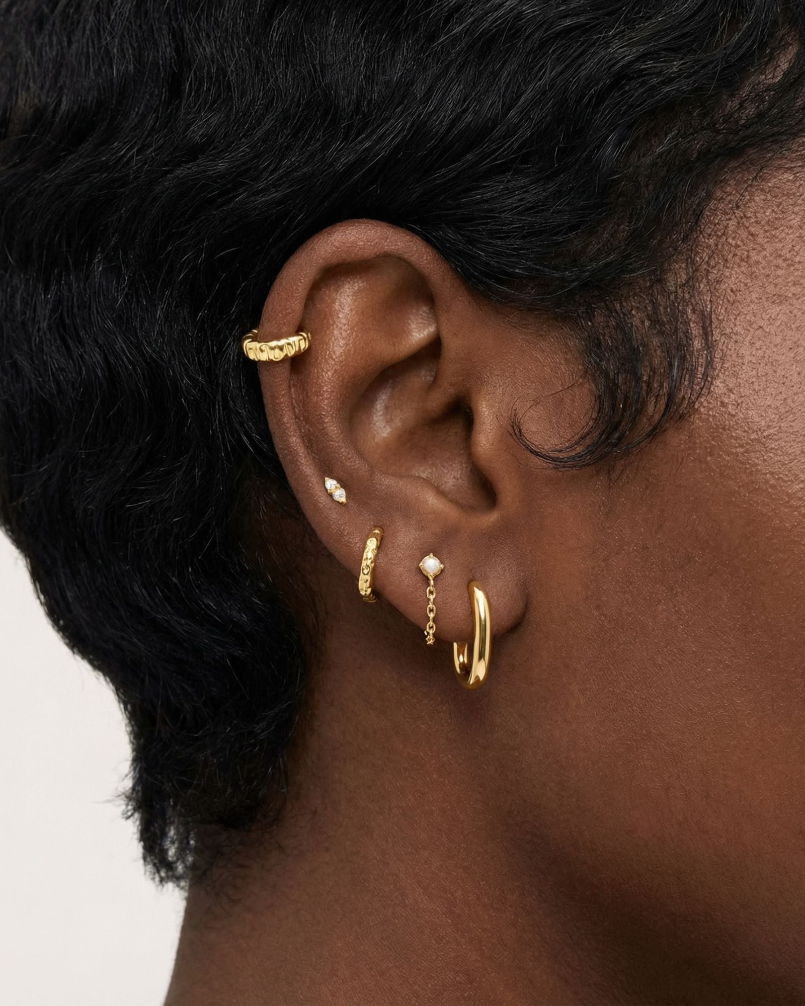 Close-up product photo of a woman’s ear with deep brown skin and natural textured hair, styled with layered gold earrings across multiple piercings. The ear features a textured gold ear cuff along the upper cartilage, a small marquise-shaped stone stud, a slim textured gold huggie, a delicate gold chain drop earring with a small round stone, and a smooth gold huggie hoop in the lobe. This image is for customers looking for versatile gold earrings that work across multiple piercings and ear placements. It he