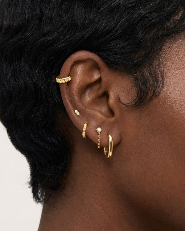 Close-up product photo of a woman’s ear with deep brown skin and natural textured hair, styled with layered gold earrings across multiple piercings. The ear features a textured gold ear cuff along the upper cartilage, a small marquise-shaped stone stud, a slim textured gold huggie, a delicate gold chain drop earring with a small round stone, and a smooth gold huggie hoop in the lobe. This image is for customers looking for versatile gold earrings that work across multiple piercings and ear placements. It he