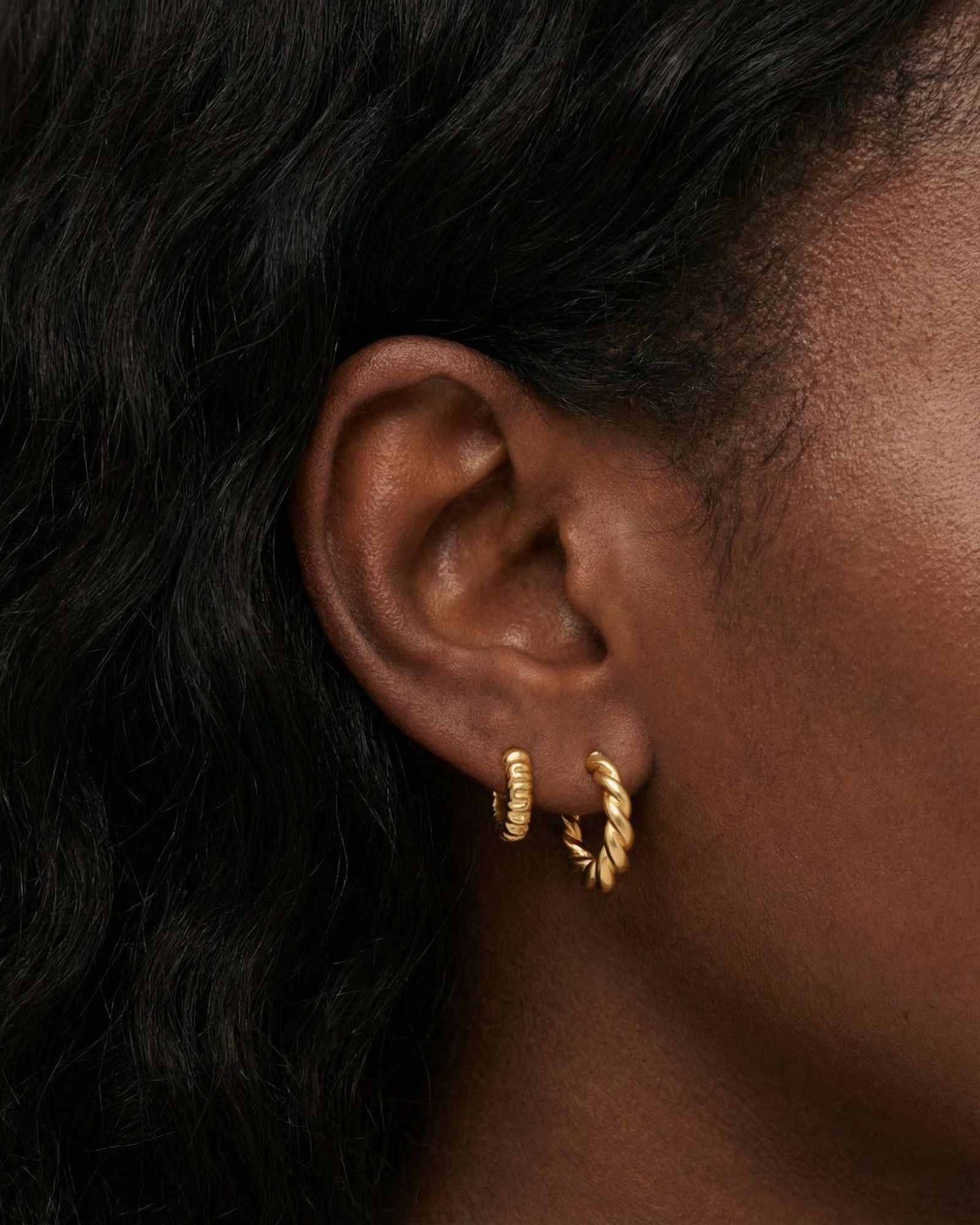 Close-up side profile of a woman’s ear wearing two small gold hoop earrings, including a twisted rope-style gold hoop and a smooth, ridged gold huggie. Designed for women who want classic gold hoop earrings for everyday wear, this image helps customers see the size, texture, and fit of layered gold hoops on the ear, making it easier to choose between subtle and statement hoop styles.