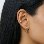Close-up product photo of a woman’s ear with warm medium skin tone and sleek dark hair, styled with minimalist gold earrings. The ear features a ribbed gold ear cuff worn on the inner cartilage and a small twisted gold huggie hoop in the lobe. This image is for customers looking for modern gold earrings that offer a clean, understated look without multiple piercings. It shows how a gold ear cuff and a simple huggie hoop can create a balanced, contemporary ear stack that adds texture and interest while remai