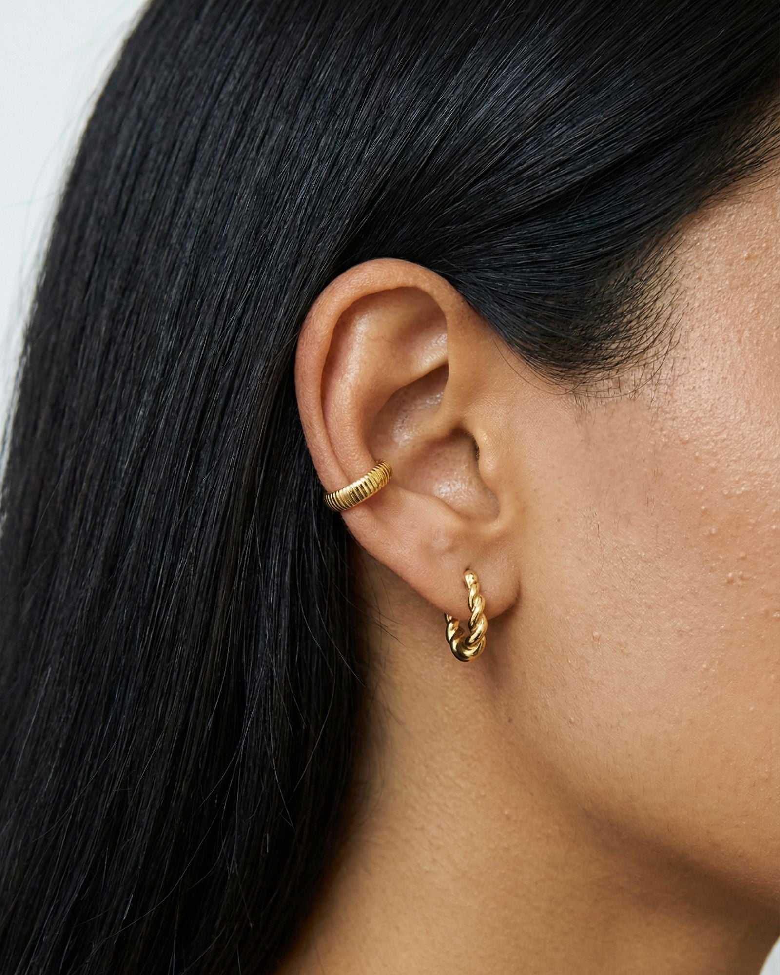 Close-up product photo of a woman’s ear with warm medium skin tone and sleek dark hair, styled with minimalist gold earrings. The ear features a ribbed gold ear cuff worn on the inner cartilage and a small twisted gold huggie hoop in the lobe. This image is for customers looking for modern gold earrings that offer a clean, understated look without multiple piercings. It shows how a gold ear cuff and a simple huggie hoop can create a balanced, contemporary ear stack that adds texture and interest while remai