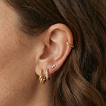 Close-up product photo of a woman’s ear with fair skin and light brown hair, styled with layered gold earrings across multiple piercings. The ear features a twisted gold huggie hoop in the first lobe piercing, a slim gold pavé huggie with clear stones in the second lobe piercing, a tiny marquise-shaped stone stud in the mid ear, and a small textured gold hoop in the upper cartilage. This image is for customers looking for refined gold earrings that combine texture and subtle sparkle for everyday wear. It sh