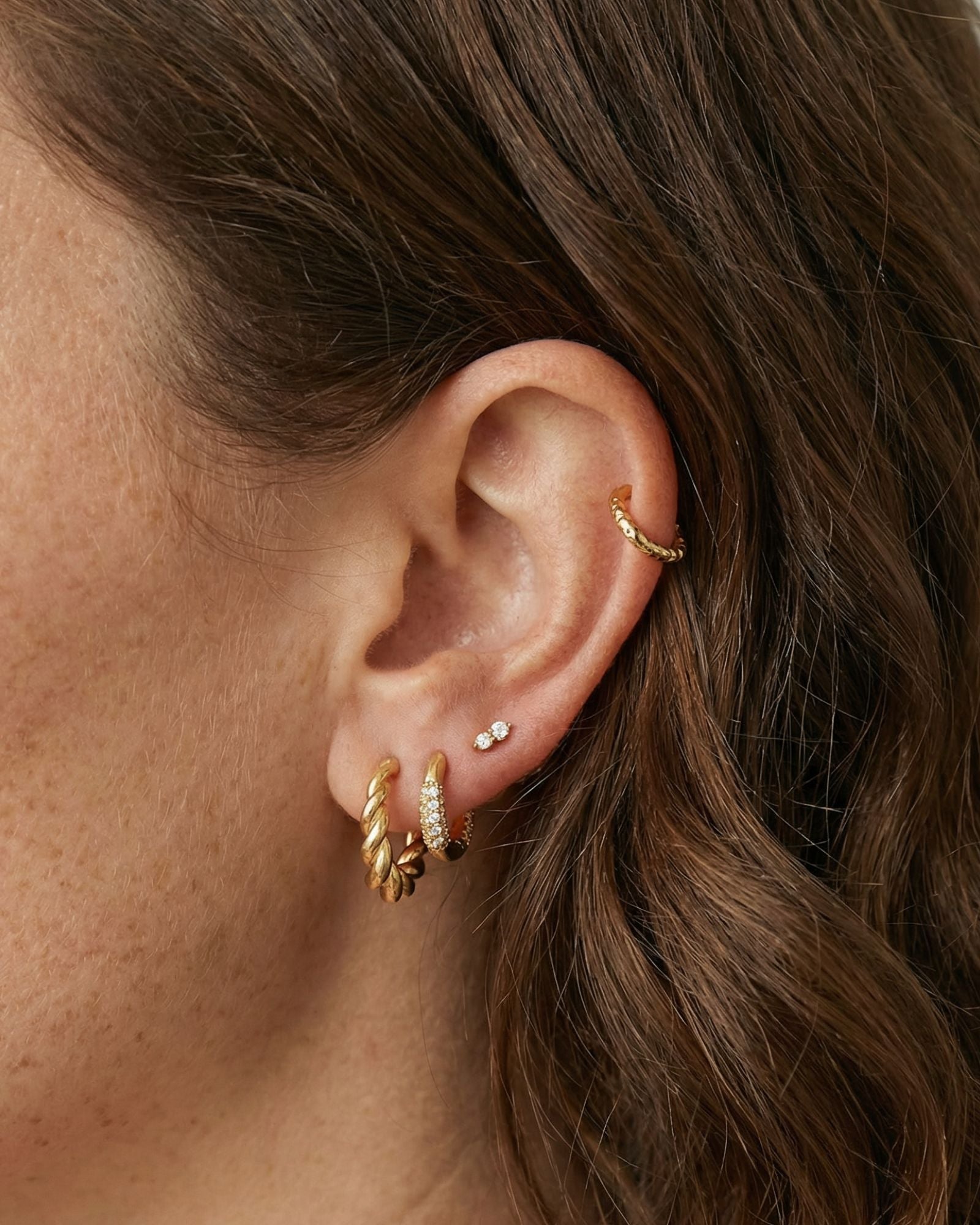 Close-up product photo of a woman’s ear with fair skin and light brown hair, styled with layered gold earrings across multiple piercings. The ear features a twisted gold huggie hoop in the first lobe piercing, a slim gold pavé huggie with clear stones in the second lobe piercing, a tiny marquise-shaped stone stud in the mid ear, and a small textured gold hoop in the upper cartilage. This image is for customers looking for refined gold earrings that combine texture and subtle sparkle for everyday wear. It sh