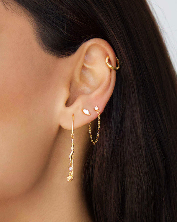 Close-up of a woman wearing the Zelia chain stud earring with marquise and round stones connected by a delicate gold chain, styled with multiple gold earrings for a layered ear stack look.
