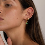 Close-up of a woman wearing small gold hoop earrings with a dangling emerald green cubic zirconia charm, styled with additional gold studs in multiple ear piercings. The image shows how the emerald charm hoops look worn on the ear, highlighting their size, movement, and layered styling potential. Designed for women seeking versatile everyday gold earrings that add subtle colour, birthstone detail, and interest to a curated ear stack without feeling heavy or overstyled.