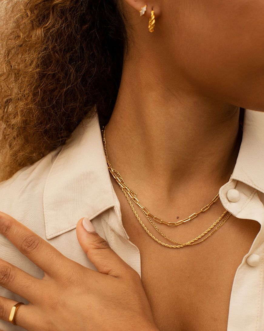 Model wearing layered 18K gold-filled chain necklaces on the collarbone, styled with a cream blouse and matching gold hoop earrings. Designed for women seeking everyday minimalist gold jewellery, this layered necklace look shows how fine gold chains add depth, balance, and effortless polish to a modern outfit without overpowering the neckline.