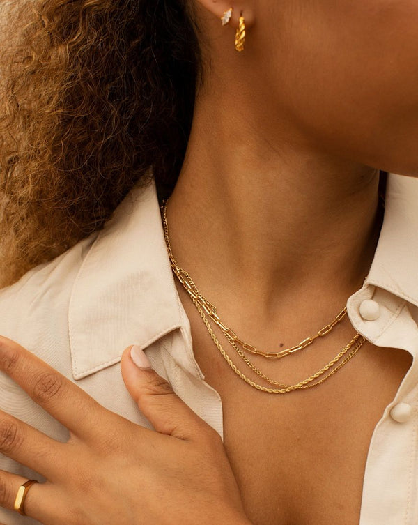 Model wearing layered 18K gold-filled chain necklaces on the collarbone, styled with a cream blouse and matching gold hoop earrings. Designed for women seeking everyday minimalist gold jewellery, this layered necklace look shows how fine gold chains add depth, balance, and effortless polish to a modern outfit without overpowering the neckline.