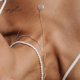 Layered gold necklaces styled on a female model lying on sand, featuring a fine gold chain with a small round coin pendant, a slim gold Cuban chain, and a white pearl strand. Worn with a minimal white swimsuit, this image is for women seeking versatile gold necklaces and pearl jewelry that work for beach, holiday, or summer styling. It helps show how to layer delicate gold chains and pearls together for a relaxed yet elevated look that transitions from swimwear to everyday outfits while maintaining a refine
