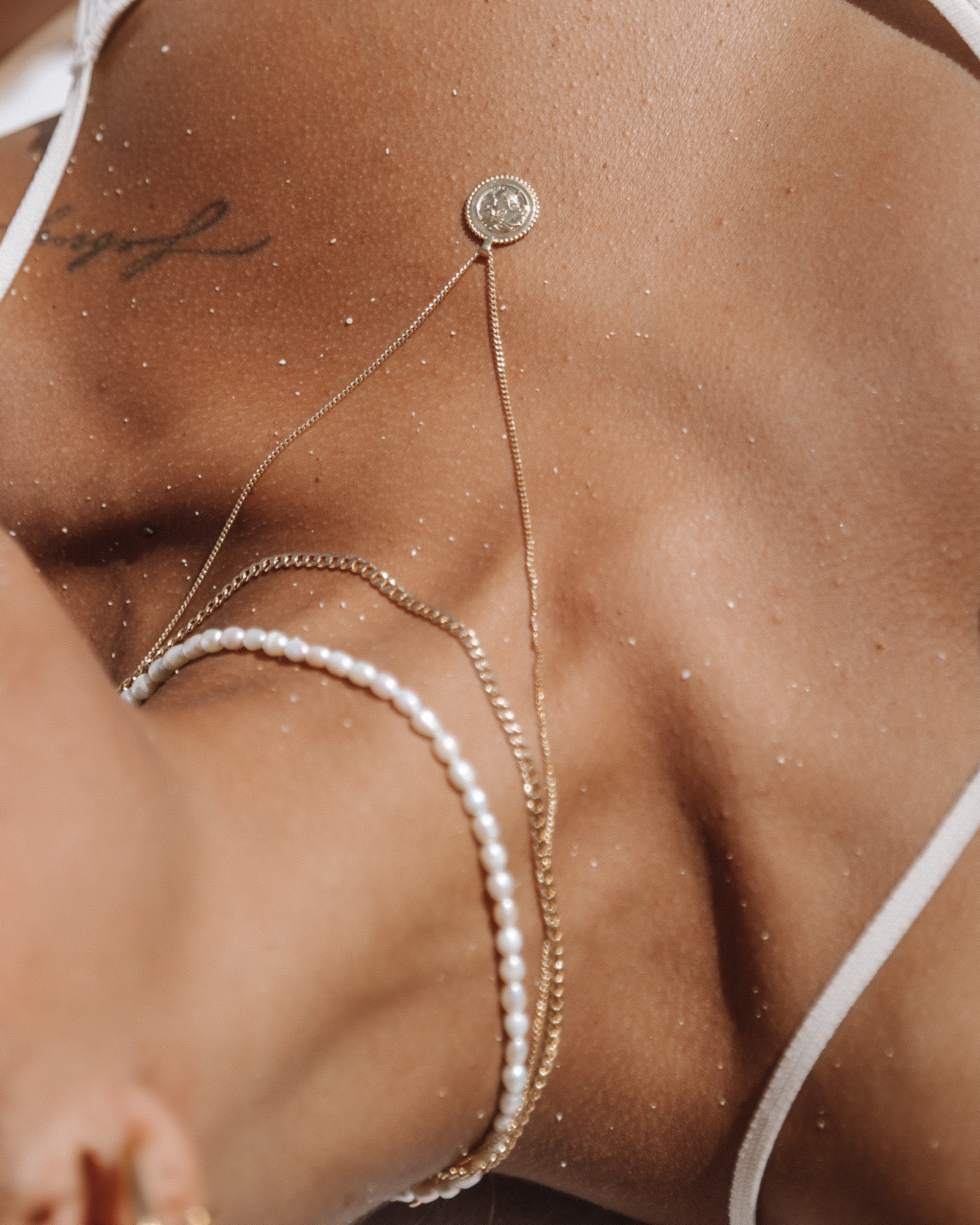 Layered gold necklaces styled on a female model lying on sand, featuring a fine gold chain with a small round coin pendant, a slim gold Cuban chain, and a white pearl strand. Worn with a minimal white swimsuit, this image is for women seeking versatile gold necklaces and pearl jewelry that work for beach, holiday, or summer styling. It helps show how to layer delicate gold chains and pearls together for a relaxed yet elevated look that transitions from swimwear to everyday outfits while maintaining a refine