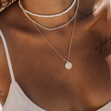 Layered gold necklaces styled on a female model wearing a white ribbed bikini top, featuring a short white pearl choker, a mid-length gold Cuban chain, and a fine gold chain with a small round coin pendant. This image is for women looking for gold necklaces and pearl jewelry suitable for summer, beach, and resort wear. It demonstrates how to layer pearl chokers with gold chains to create a balanced, effortless necklace stack that elevates swimwear and warm-weather outfits while maintaining a clean, modern l