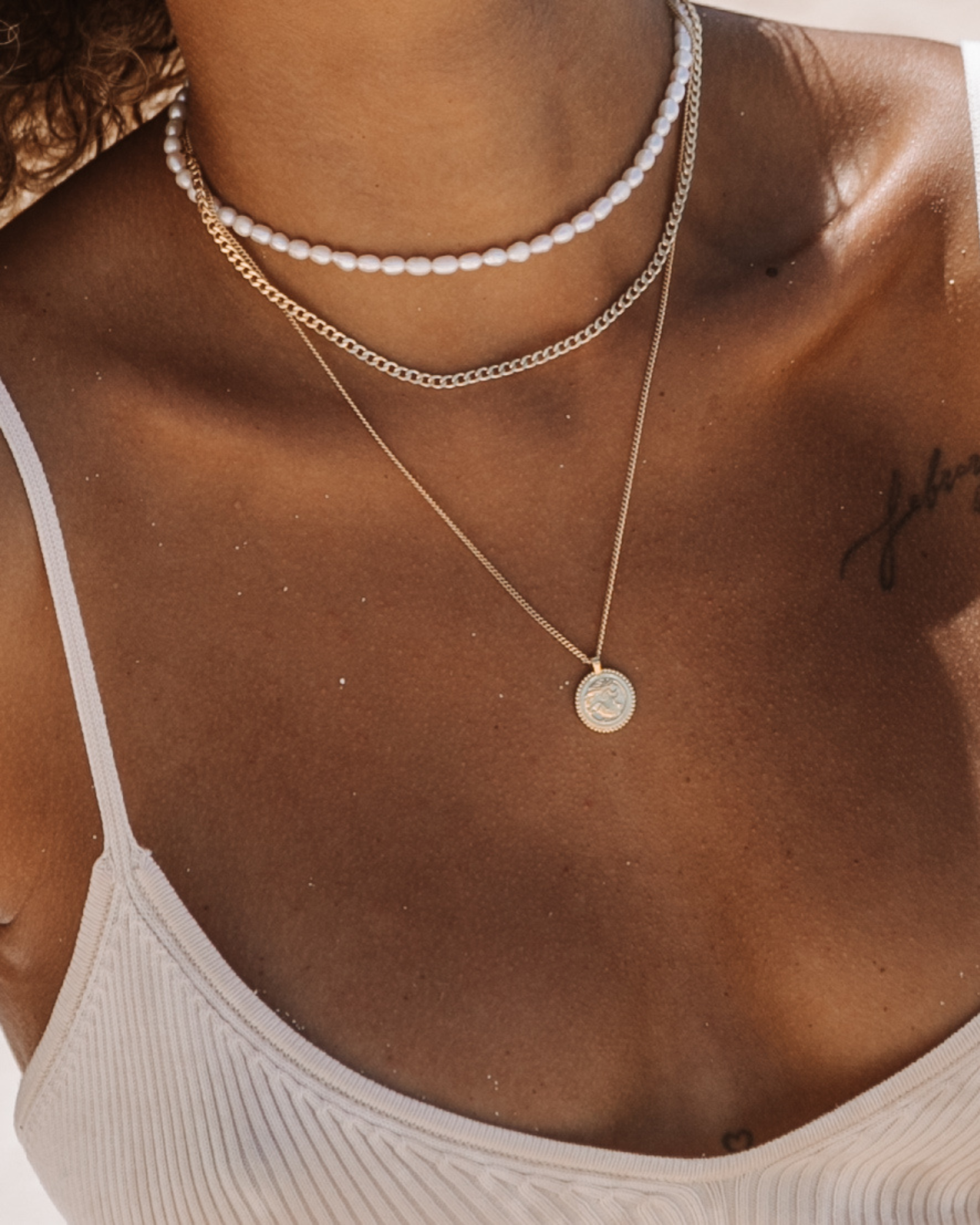 Layered gold necklaces styled on a female model wearing a white ribbed bikini top, featuring a short white pearl choker, a mid-length gold Cuban chain, and a fine gold chain with a small round coin pendant. This image is for women looking for gold necklaces and pearl jewelry suitable for summer, beach, and resort wear. It demonstrates how to layer pearl chokers with gold chains to create a balanced, effortless necklace stack that elevates swimwear and warm-weather outfits while maintaining a clean, modern l