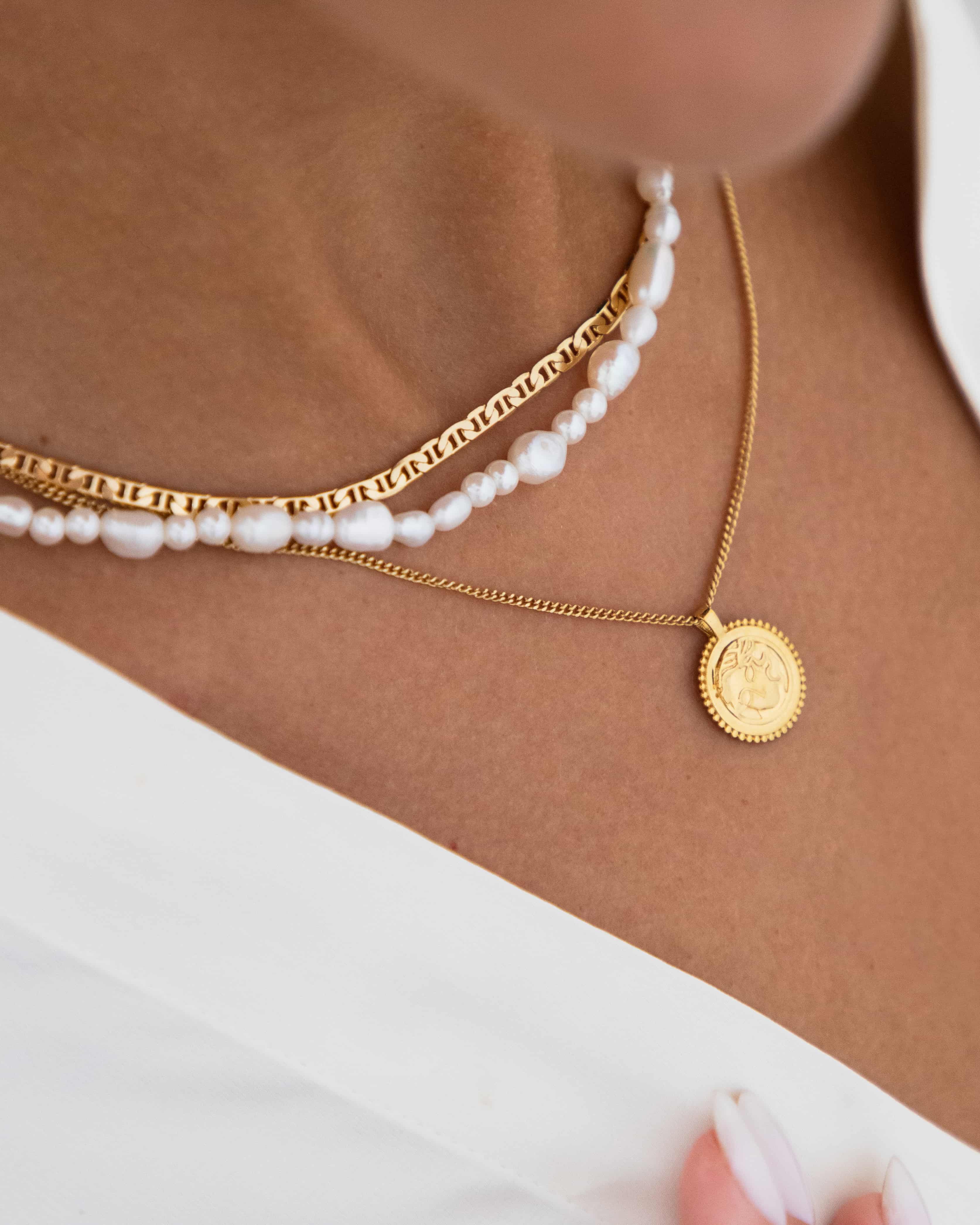 Layered gold necklaces styled on the neck, featuring a slim flat-link gold chain, a freshwater pearl necklace, and a delicate gold coin pendant on a fine chain. Designed for women looking for elegant layered gold necklaces, pearl and gold combinations, and easy everyday styling, helping create a balanced, polished jewellery stack without needing multiple statement pieces.