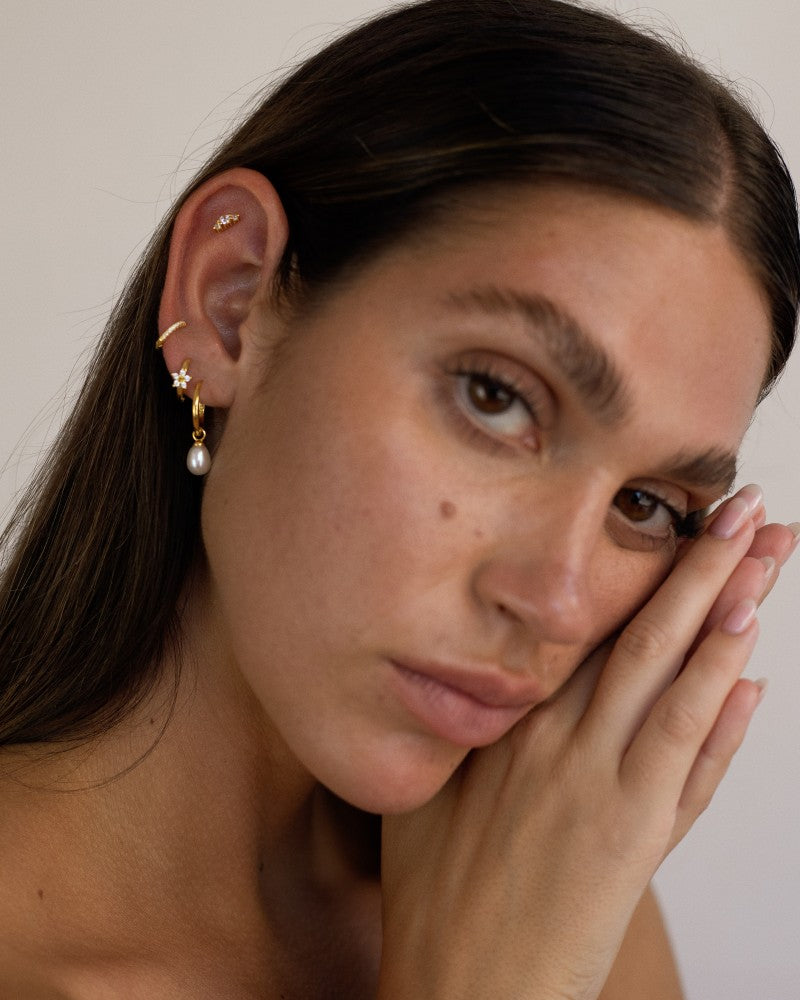 Close-up portrait of a woman wearing gold pearl hoop earrings with a single dangling freshwater pearl, styled with a small gold star stud, a slim gold ear cuff, and a delicate crystal cartilage stud. Designed for women who love minimalist gold jewellery and curated ear styling, this image shows how pearl hoop earrings can be layered for a soft, elegant look that works for everyday wear, weddings, and special occasions. 