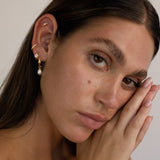 Close-up portrait of a woman wearing gold pearl hoop earrings with a single dangling freshwater pearl, styled with a small gold star stud, a slim gold ear cuff, and a delicate crystal cartilage stud. Designed for women who love minimalist gold jewellery and curated ear styling, this image shows how pearl hoop earrings can be layered for a soft, elegant look that works for everyday wear, weddings, and special occasions. 