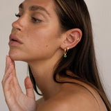 Side profile of a woman wearing small gold hoop earrings with a dangling emerald green cubic zirconia charm, styled with a tiny gold huggie and a minimal crystal stud in the upper ear. The image shows how the emerald charm hoop sits on the lobe and moves naturally when worn, highlighting scale, layering potential, and everyday elegance. Designed for women looking for versatile gold hoop earrings with a subtle pop of green, suitable for daily wear, curated ear stacks, and refined, modern jewellery styling.