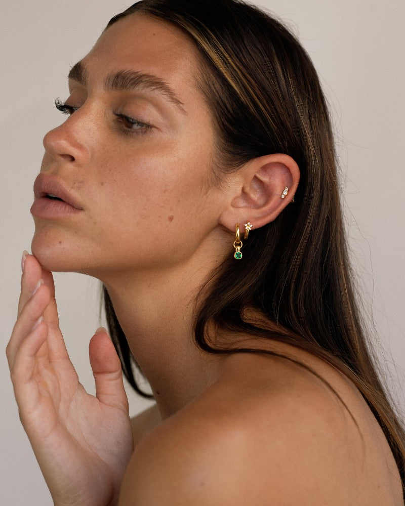 Side profile portrait of a woman wearing a curated gold ear stack featuring a flat back cartilage stud with cubic zirconia, a small gold huggie in the lobe, and a gold hoop with a green gemstone drop. The image demonstrates how flat back earrings and cartilage studs can be layered for a refined, everyday ear stack, ideal for those seeking comfortable, secure jewellery for sensitive ears and long-term wear.