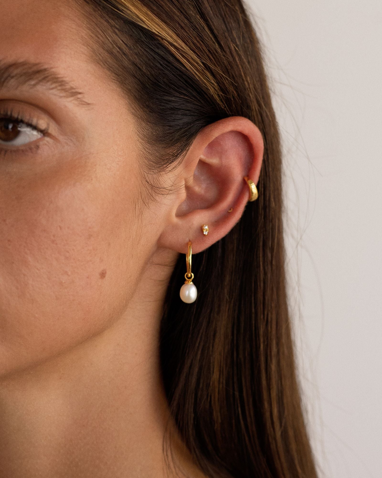 Close-up of a woman’s ear wearing a small gold hoop with a freshwater pearl drop, a dainty gold stud, and a slim gold ear cuff, styled to show minimalist gold jewellery and pearl drop earrings for everyday wear, ear stack layering, and refined, comfortable styling for sensitive ears.