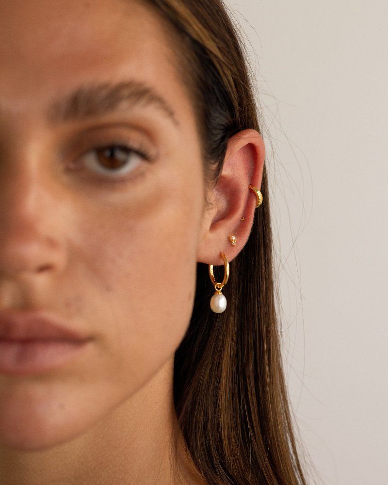 Close-up portrait of a woman wearing gold pearl hoop earrings with a dangling freshwater pearl, paired with a small gold stud and a gold ear cuff on the cartilage. Designed for women who enjoy modern, minimalist jewellery and curated ear stacks, this image shows how pearl hoop earrings can be styled with studs and cuffs for a balanced everyday look or elevated occasion wear. Ideal for those seeking versatile gold pearl earrings that add softness